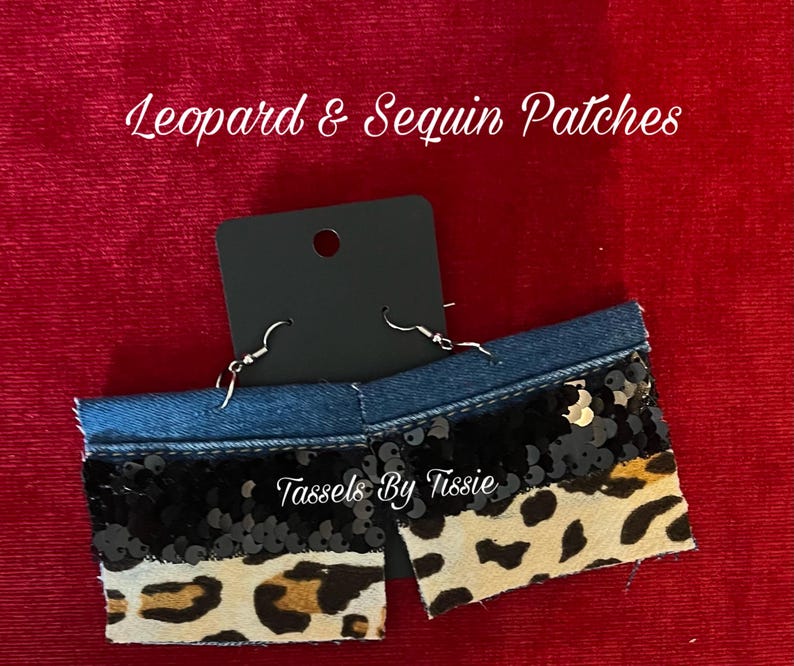 May include: A pair of denim earrings with black sequins and a leopard print fabric. The earrings are handmade and have a silver hook closure. The earrings are labeled "Tassels By Tisse".