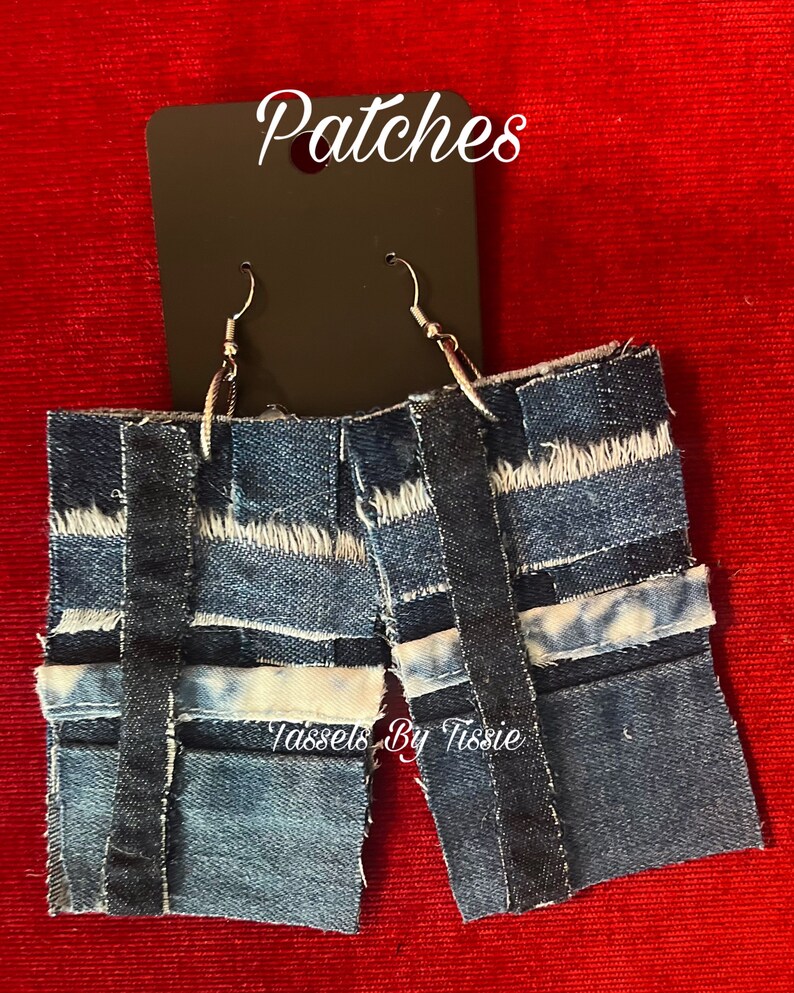 May include: A pair of denim earrings with a patchwork design. The earrings are blue and have a frayed edge. The earrings are labeled "Patches" and "Tassels By Tisse".