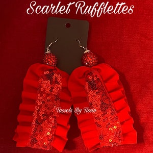 May include: A pair of red tassel earrings with a sequined center and a rhinestone ball at the top. The earrings are displayed on a black card against a red background. The text "Scarlet Rufflettes" is at the top.