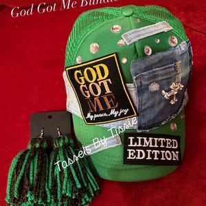 May include: A green trucker hat featuring a denim patch, a black patch with the text GOD GOT ME My peace, My joy, a LIMITED EDITION patch, and scattered rhinestones. Beside it are two green and black yarn tassel earrings on a black card.