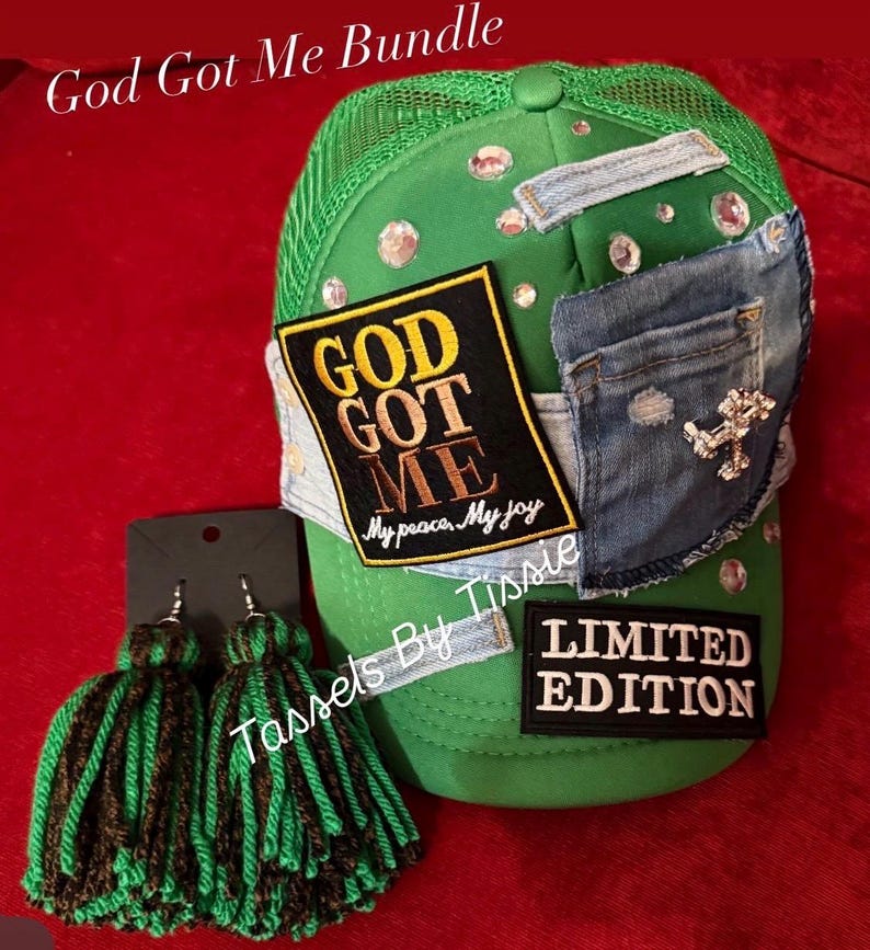 Może przedstawiać: Zielona czapka typu trucker z dżinsową naszywką, czarną naszywką z napisem GOD GOT ME My peace, My joy, naszywką LIMITED EDITION oraz rozrzuconymi cyrkoniami. Obok znajdują się dwa kolczyki z zielono-czarnymi frędzlami z wł&oacute;czki na czarnej karcie.