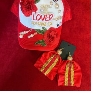 Może przedstawiać: Czerwono-biała czapka typu trucker z haftowanymi czerwonymi r&oacute;żami, naszywką z napisem "ORIGINAL", "LOVED" i "ROMANS 5:8", ozdobami z pereł i cyrkonii oraz parą pasujących czerwonych kolczyk&oacute;w z frędzlami ze złotymi akcentami brokatowymi.