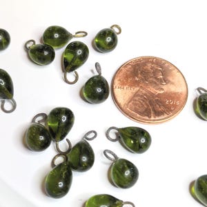May include: A collection of small, olive-green glass teardrop-shaped beads with attached metal loops. A copper penny is included for size reference. The beads are scattered on a white surface.