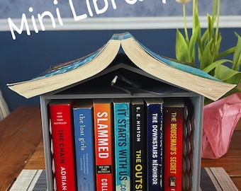 Może przedstawiać: Miniaturowa biblioteczka wykonana z szarego drewna z dachem w kształcie książki. Biblioteczka jest wypełniona książkami w miękkiej oprawie z tytułami takimi jak "The Chain", "The Lost Girls", "Slammed", "It Starts With Us", "The Outsiders", "The Downstairs Neighbor" i "The Housemaid's Secret".