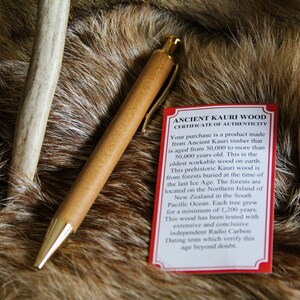 May include: A wooden pen with gold accents on a brown fur background. A red card with the text "Ancient Kauri Wood Certificate of Authenticity" is visible. The card explains that the wood is over 30,000 years old and is from New Zealand.