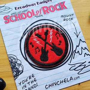 May include: A patch with the text "SCHOOL OF ROCK" and a design of two guitars inside a red circle. The patch is on a lined paper with the text "YOU'RE IN THE BAND!" and "CHINCHELA.com". Pencils, scissors, and a crumpled paper are also visible.