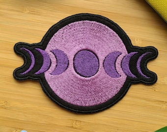 May include: Purple and black embroidered patch with three crescent moons and a circle in the center. The patch is on a wooden surface with colorful pens and a tie-dye fabric.