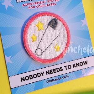 May include: A circular embroidered patch with a pink border featuring a safety pin and yellow stars on a white background. The patch is mounted on a blue card labeled COSPLAY BADGES ACHIEVEMENT SYSTEM FOR COSPLAYERS and NOBODY NEEDS TO KNOW.
