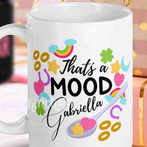 Puede incluir: Taza de cerámica blanca con un diseño negro y colorido. La taza tiene el texto "That's a MOOD Gabriella" con un arcoíris, estrellas, corazones y tréboles.