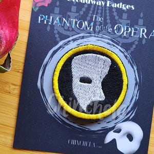Op de afbeelding: Een Broadway Badges-kaart met een Phantom of the Opera-patch. De patch heeft een wit maskerontwerp met een gele rand op een zwarte achtergrond. Een wit masker staat onder de patch. Een roos staat in de linkerbovenhoek.