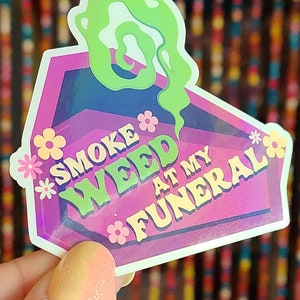 May include: A pink and purple holographic sticker with a green smoke design. The sticker says "Smoke Weed at My Funeral".