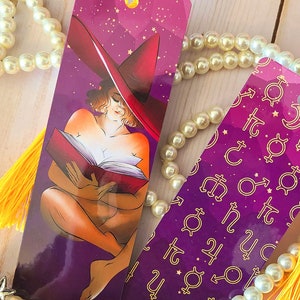 May include: Two purple and gold bookmarks with yellow tassels. One bookmark features a woman in a red hat reading a book. The other bookmark has a repeating pattern of astrological symbols.