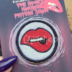May include: A patch featuring a red, open-mouthed design with a tongue sticking out, surrounded by a white border and a black outline. The patch is on a card with the text "Broadway Badges" and "The Rocky Horror Picture Show". The website address CHINCHELA.com is also visible.