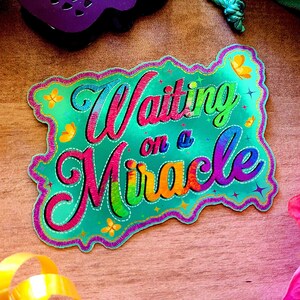May include: A colorful, glittery sticker with the text "Waiting on a Miracle" in a rainbow font. The sticker has a green background with a glittery finish.