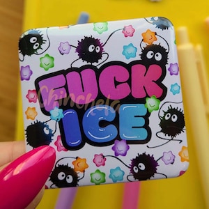 May include: A white square pin with the text "FUCK ICE" in pink and blue, surrounded by cartoonish black creatures and colorful stars. A matching sticker is also visible. The pin is held in front of a yellow notebook.