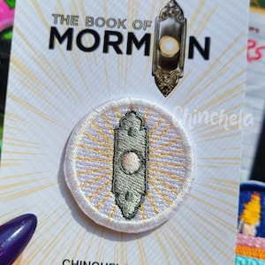 Könnte beinhalten: Ein runder gestickter Aufnäher mit einem grauen Türklingel-Symbol und gelben, strahlenförmigen Linien auf weißem Grund. Der Aufnäher ist auf einer weißen Karte mit dem Text THE BOOK OF MORMON und CHINCHELA.COM befestigt.
