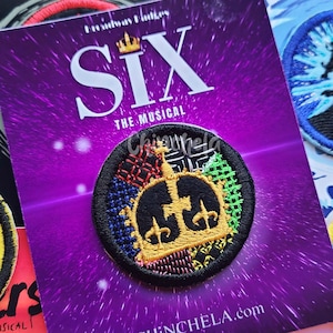 May include: A circular embroidered patch featuring a gold crown design on a multi-colored patterned background, mounted on a purple card labeled SIX THE MUSICAL and CHINCHELA.com. The patch has a black border and is surrounded by other musical theater patches.