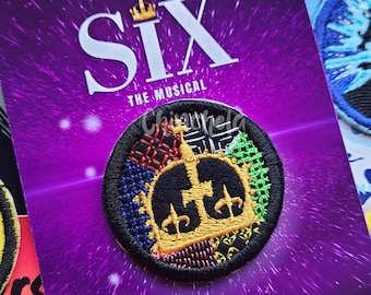 May include: A circular embroidered patch featuring a gold crown design on a multi-colored patterned background, mounted on a purple card labeled SIX THE MUSICAL and CHINCHELA.com. The patch has a black border and is surrounded by other musical theater patches.