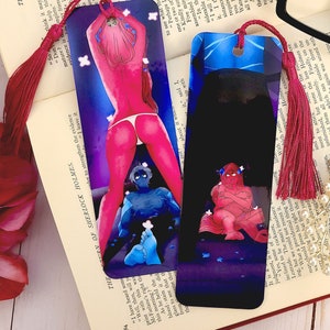 May include: Two colorful bookmarks with a dark blue and purple background. One bookmark features a woman with pink hair and a blue figure in the background. The other bookmark features a red figure with a blue background. Both bookmarks have a red tassel.