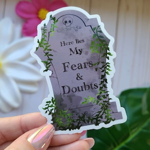 May include: A gray tombstone sticker with green vines growing around it. The tombstone reads "Here lies My Fears & Doubts".