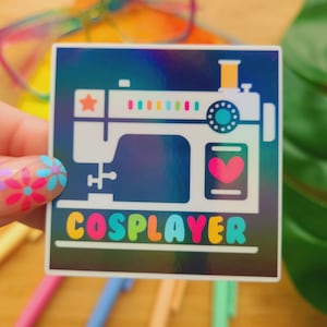 May include: A square sticker with a holographic background features a white sewing machine illustration. The word "COSPLAYER" is written in colorful letters below the machine. A pink heart is visible on the machine.