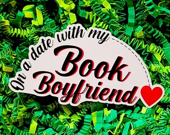 May include: A white sticker with red and black text that says "On a date with my Book Boyfriend" with a red heart on the right side. The sticker is on a green and yellow shredded paper background.