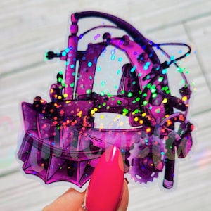 May include: A transparent sticker featuring a purple, steampunk-inspired design with gears, arches, and decorative elements. The sticker has a glittery, holographic effect, with a hand holding it up.