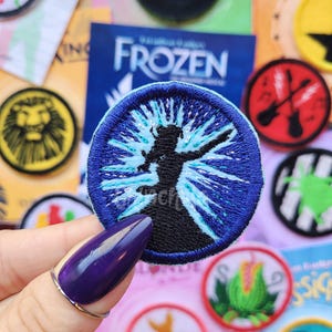 May include: A circular embroidered patch featuring a black silhouette of a figure with one arm raised, surrounded by a bright blue starburst design with silver metallic thread accents, all enclosed within a dark blue stitched border.