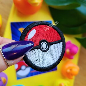 May include: A circular embroidered patch featuring a red and white Poké Ball design with a black border and a central button detail. The patch has a white highlight on the upper red section and is stitched with dense thread in a classic round shape.