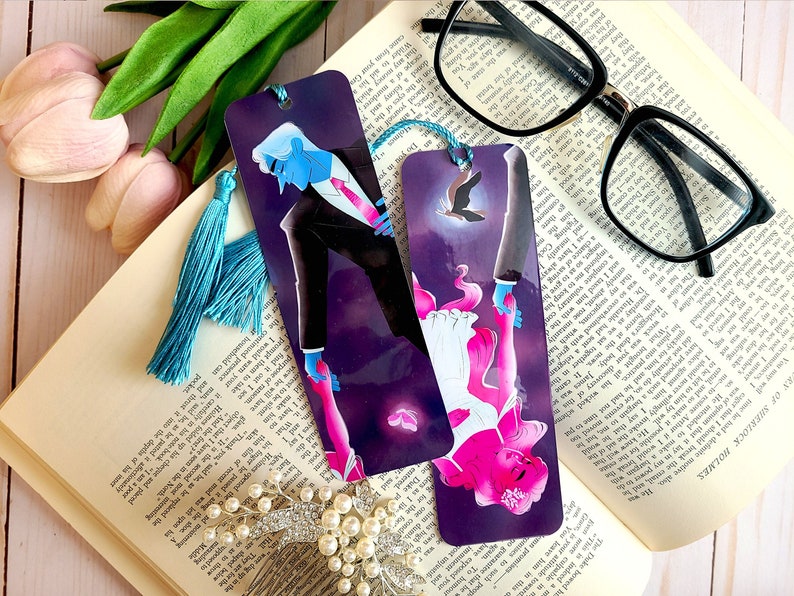 May include: Two bookmarks with a purple background and a couple holding hands. The man is wearing a black suit and the woman is wearing a white dress. The bookmark on the left has a blue tassel and the bookmark on the right has a black tassel.