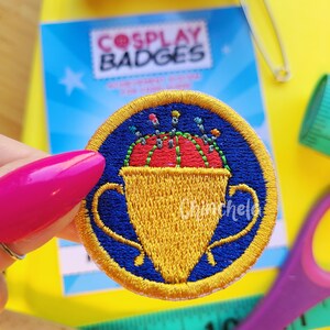 May include: A circular embroidered patch featuring a gold trophy cup filled with a red pincushion containing colorful sewing pins, set against a dark blue background with a gold border. The word Chinchela is stitched in white on the right side.