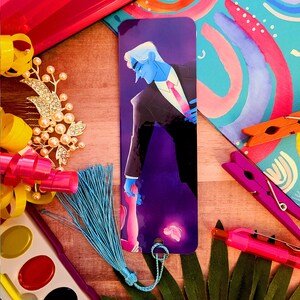 May include: A purple and black bookmark with a blue-haired man in a suit holding a hand of a person with a blue tassel. The bookmark has a butterfly design and a string to attach it to a book.