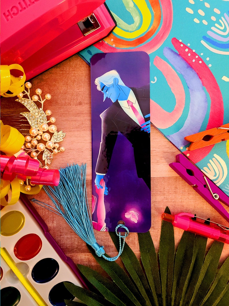 May include: A purple and black bookmark with a blue-haired man in a suit holding a hand of a person with a blue tassel. The bookmark has a butterfly design and a string to attach it to a book.