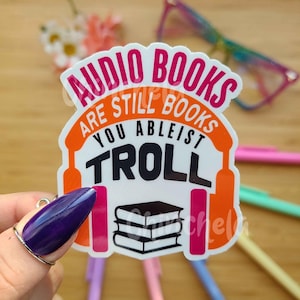 May include: A white sticker with orange and pink text that reads "Audio Books Are Still Books You Ableist Troll". The sticker features a black and white illustration of a stack of books with headphones on either side.