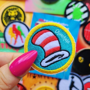 May include: A circular embroidered patch featuring a red and white striped hat on a teal background with a yellow border. The text Chinchela is stitched in white script on the right side of the patch. The design is centered on a round fabric base.