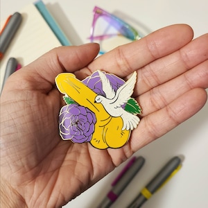 May include: A gold-tone enamel pin featuring a yellow banana with a white dove perched on top. The banana is surrounded by purple flowers with green leaves.