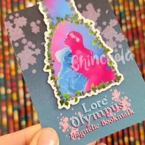 May include: A blue and pink magnetic bookmark with a floral design and the text "Lore Olympus Magnetic Bookmark".