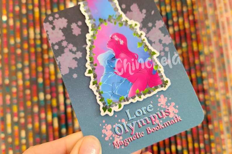 May include: A blue and pink magnetic bookmark with a floral design and the text "Lore Olympus Magnetic Bookmark".