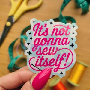 May include: A sticker with the text "It's not gonna sew itself!" in pink, with a needle and thread design. The sticker is held by a person with a pink manicure, with sewing supplies in the background.