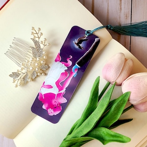 May include: A bookmark with a purple background featuring a woman with pink hair holding hands with a man in a suit. A bird flies above them. The bookmark has a teal tassel.
