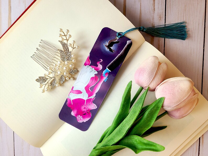 May include: A bookmark with a purple background featuring a woman with pink hair holding hands with a man in a suit. A bird flies above them. The bookmark has a teal tassel.
