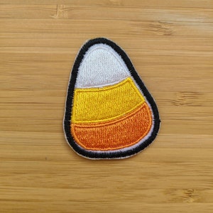 Może przedstawiać: Naszywka z motywem kukurydzy cukrowej, wyszywana czarnymi i białymi nićmi, z pomarańczowymi, żółtymi i białymi paskami.
