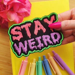 Puede incluir: Un parche de plancha negro con bordado verde, rosa y morado que dice "STAY WEIRD".