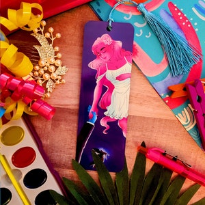 May include: A purple bookmark with a blue tassel. The bookmark features a pink-haired woman with a white dress and a black figure reaching towards her. The background is a dark purple with a glowing blue flower.