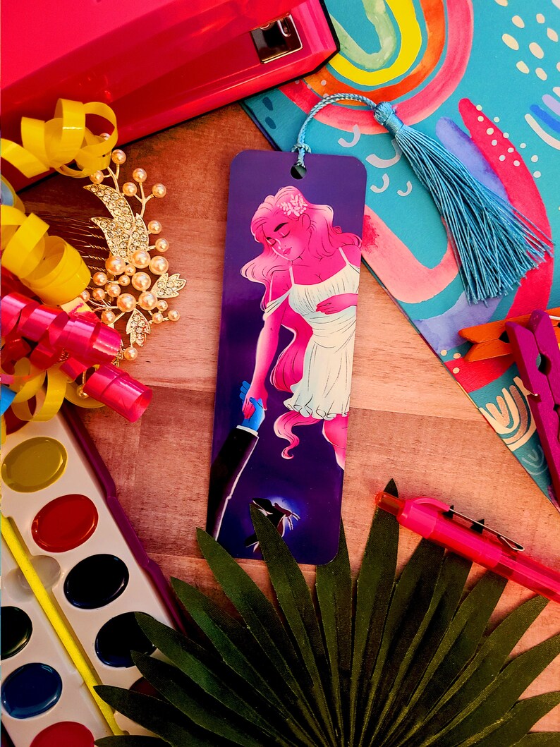 May include: A purple bookmark with a blue tassel. The bookmark features a pink-haired woman with a white dress and a black figure reaching towards her. The background is a dark purple with a glowing blue flower.