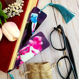 May include: Two bookmarks featuring illustrations of a man and a woman. The bookmark on the left features a man in a suit with a blue tie. The bookmark on the right features a woman with pink hair and a white dress. Both bookmarks have a teal tassel.