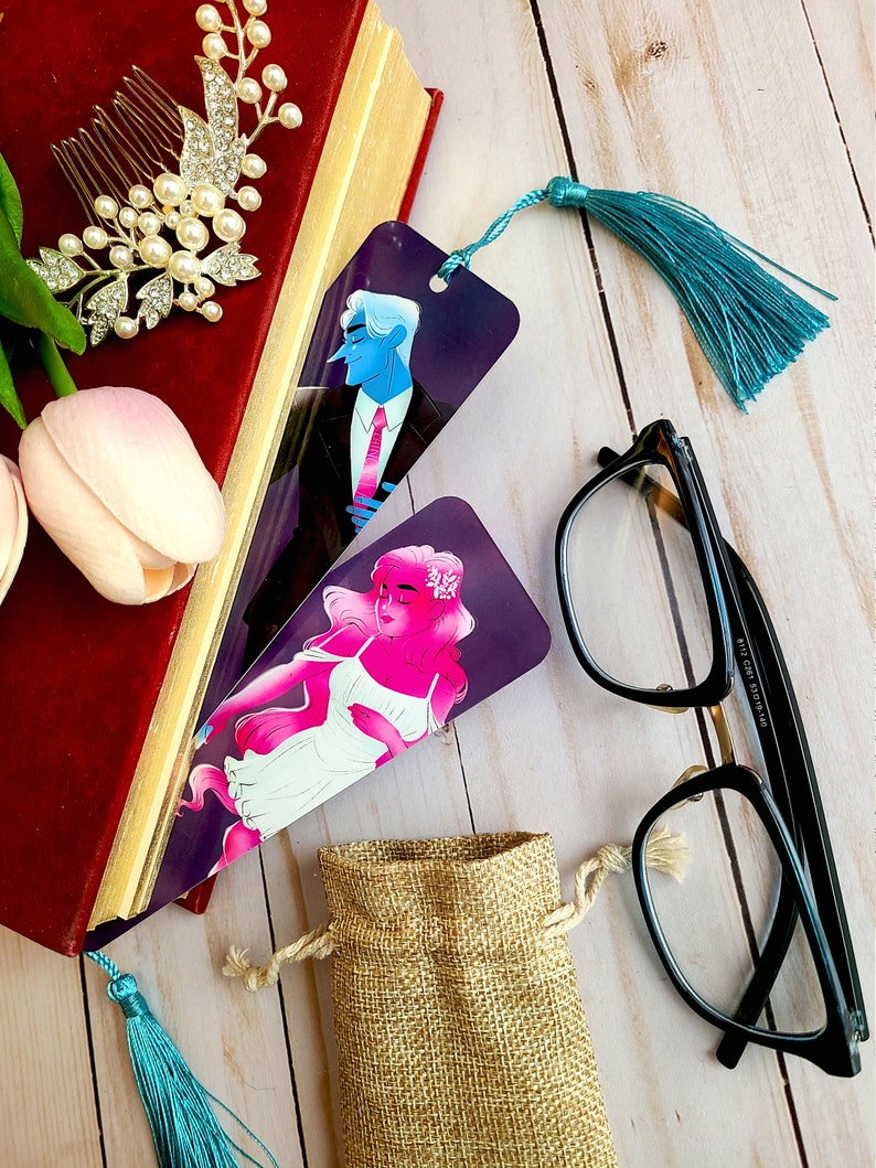 May include: Two bookmarks featuring illustrations of a man and a woman. The bookmark on the left features a man in a suit with a blue tie. The bookmark on the right features a woman with pink hair and a white dress. Both bookmarks have a teal tassel.