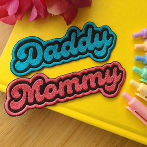 May include: Two iron-on patches with the words "Daddy" and "Mommy" in a retro font. The "Daddy" patch is blue and the "Mommy" patch is pink.