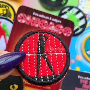 Puede incluir: Un parche bordado circular con fondo rojo, líneas de costura verticales blancas y la silueta negra de una persona en una pose. El parche tiene un borde negro grueso y se sostiene frente a una tarjeta roja con el texto CHICAGO.