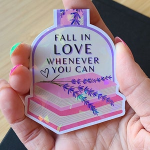 May include: A pink and purple holographic bookmark with a stack of books and lavender flowers. The bookmark has the text "Fall in Love Whenever You Can" with a heart symbol.